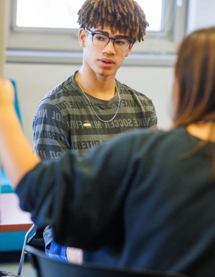 A student looks at a whiteboard as another student presents from it.